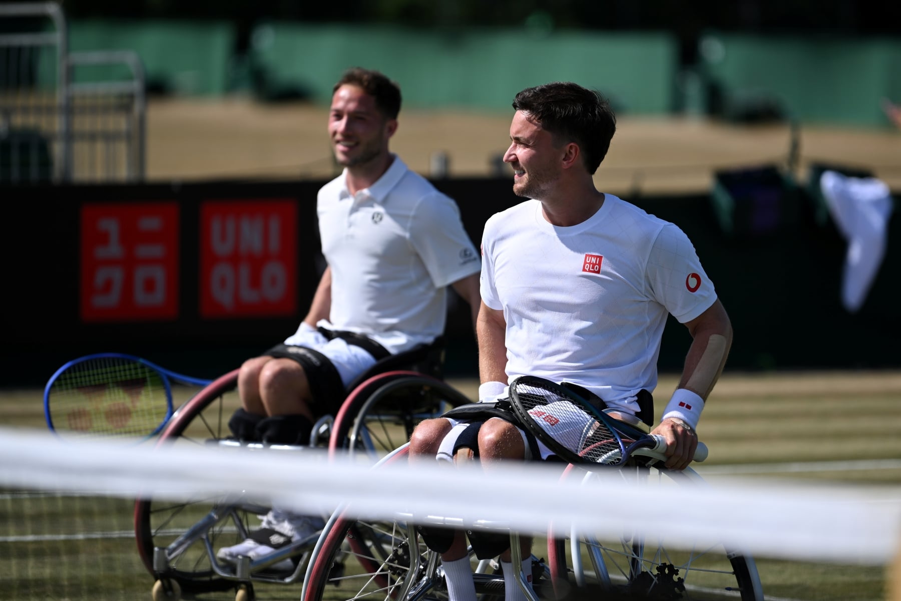 Two men in wheelchairs wearing white t-shirts and shorts and holding tennis rackets smile as they wheel themselves towards a tennis net