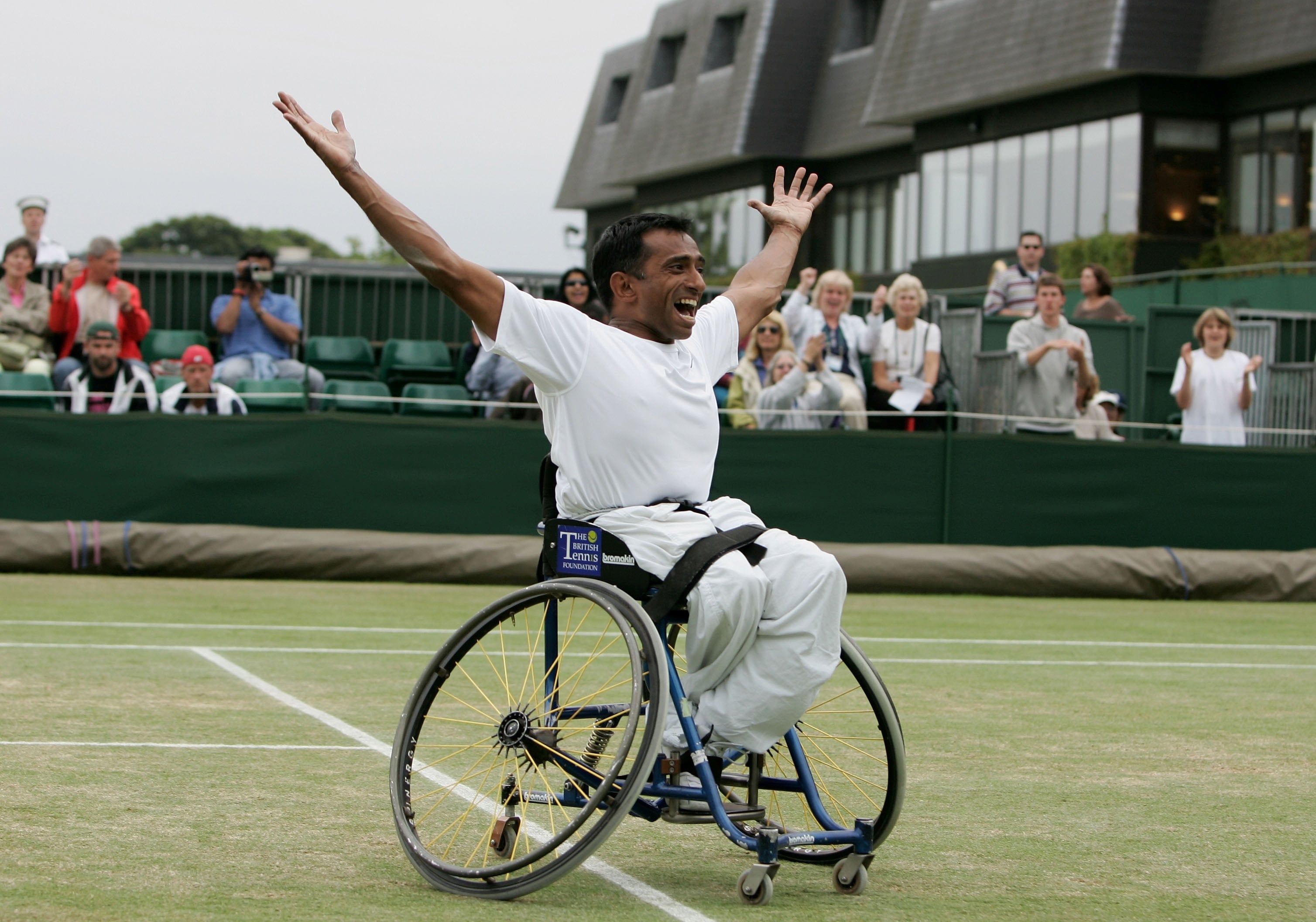 A man in a wheelchair wearing all-white clothing whilst on a grass tennis court throws his hands in the air in celebration
