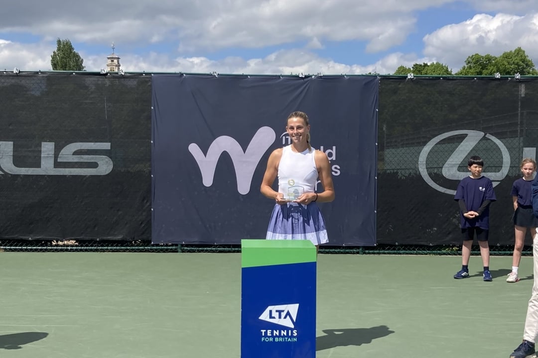 A woman wearing a white vest top and a lilac skirt holds a glass trophy, standing behind a blue pedestal with the words, 'LTA, Tennis for Britain' on it