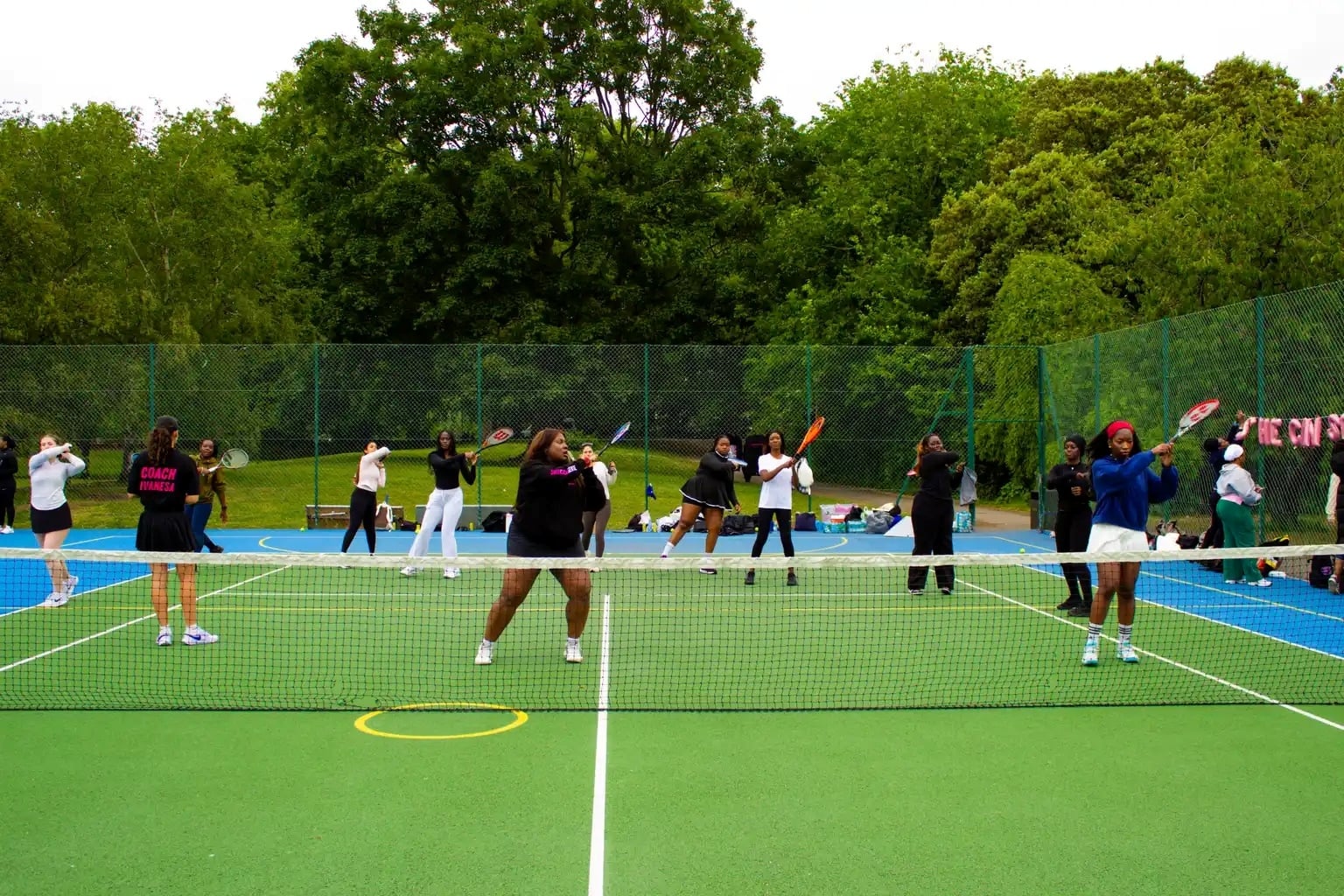 A group of women stand holding tennis rackets, on a tennis court that is green with blue outside borders. In the background, large trees coverage in green foliage are visible.