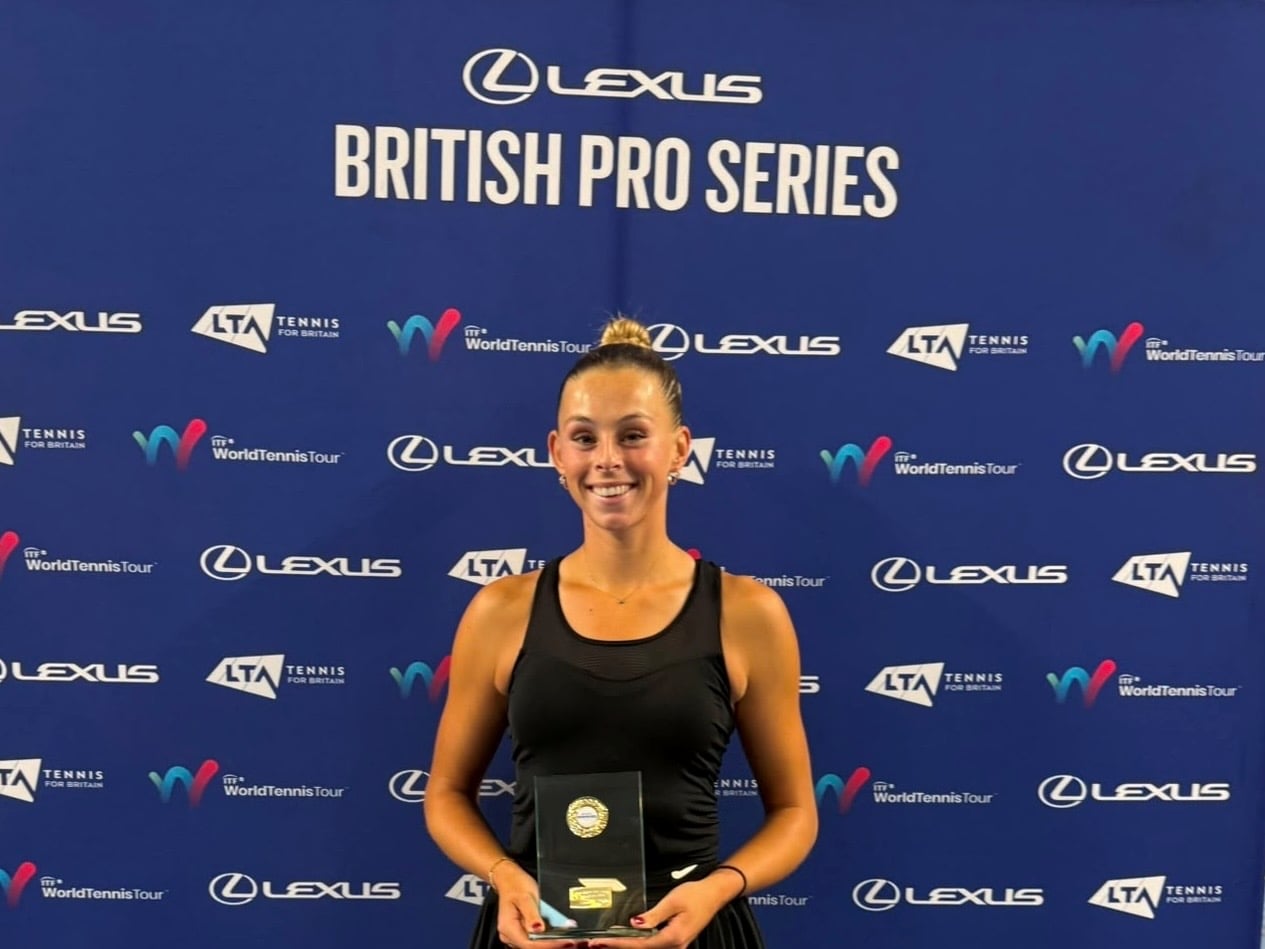 A woman wearing a black vest top satnds smiling and holding a glass trophy. Behind her is a blue backdrop featuring a variety of sponsor logos, with the words Lexus British Pro Series at the top