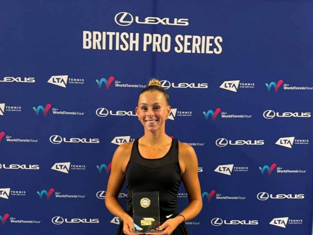 A woman wearing a black vest top satnds smiling and holding a glass trophy. Behind her is a blue backdrop featuring a variety of sponsor logos, with the words Lexus British Pro Series at the top