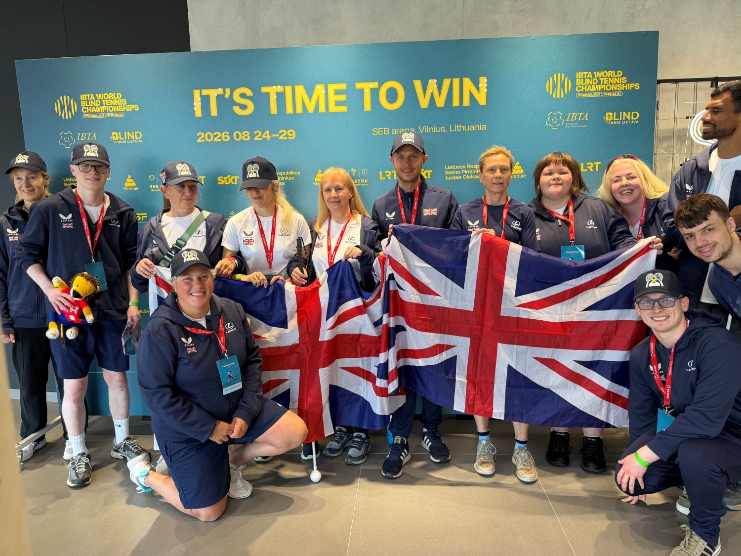 A group of people wearing dark blue sports kit stand holding two unio flags side by side. Behind the group is a light blue board that displays the words 'It's time to win'