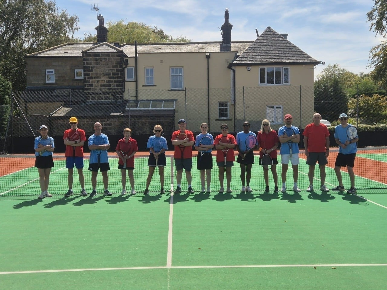 A group of people stand on a tennis court holding tennis rackets. They are stood in a line and alternate wearing light blue and red t-shirts