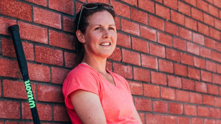 Elly Shearman leaning against a brick wall smiling with Wilson tennis racket next to her