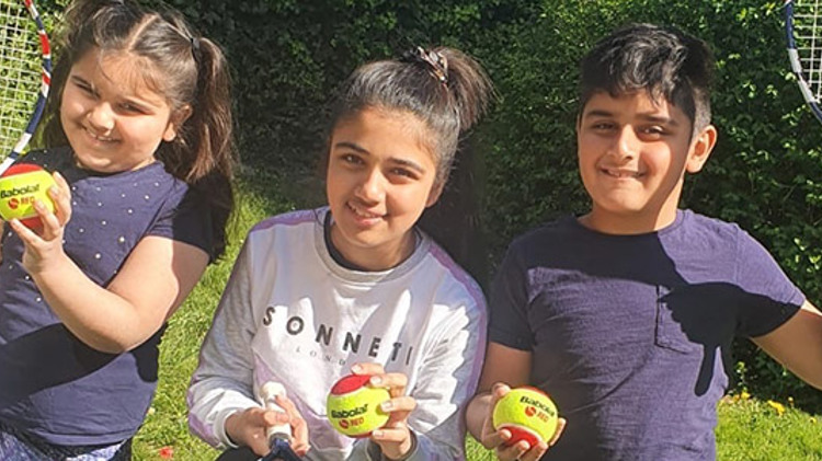 three kids smiling all holding a yellow and red tennis ball and two with tennis rackets in the air